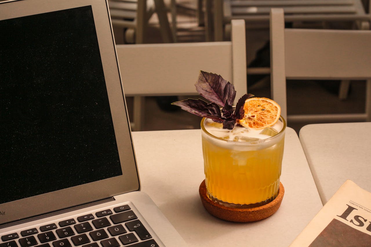 pexels-photo-27524197 A refreshing citrus cocktail garnished with basil beside a laptop and newspaper, perfect for a casual work setting.